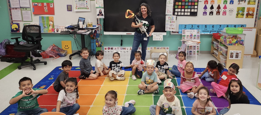 A classroom with a teacher standing in front of a colorful rug where young children sit.