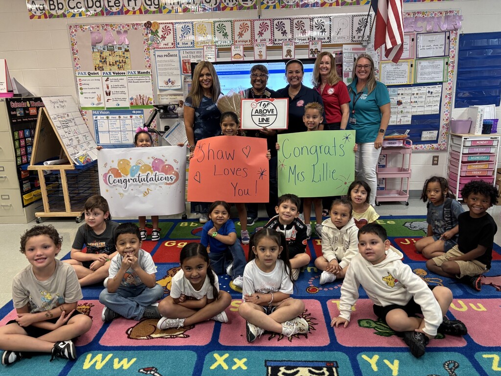 Several children and adults pose for a picture in a classroom with a rug. Adults hold signs.