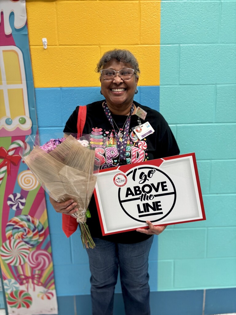 A woman holding flowers and a sign reading "I go above the line" in front of a colorful wall.