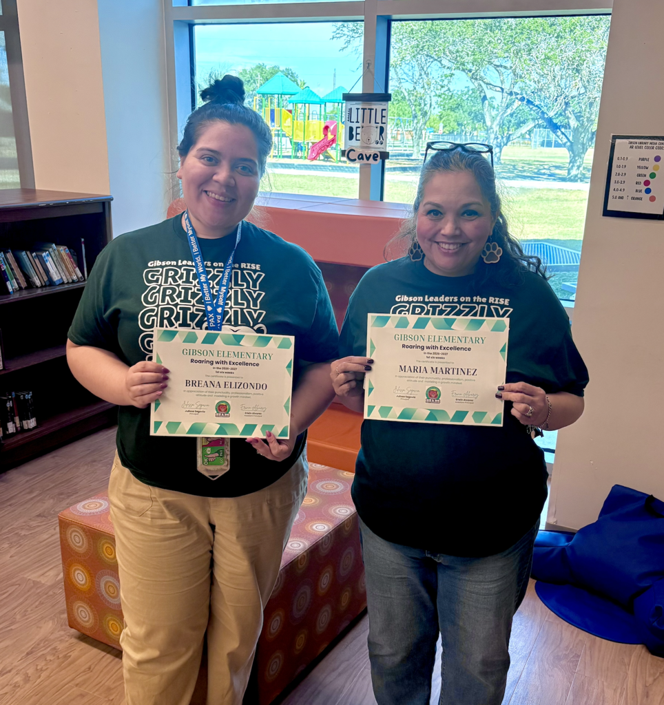 Two women stand with certificates in hand. One wears a green shirt with "Critically Growing" text. Background includes a window, playground, and shelf.