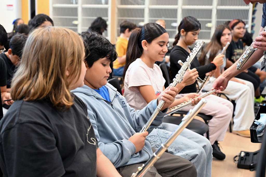 Students seated in a classroom hold flutes. One student holds a flute while a teacher assists.