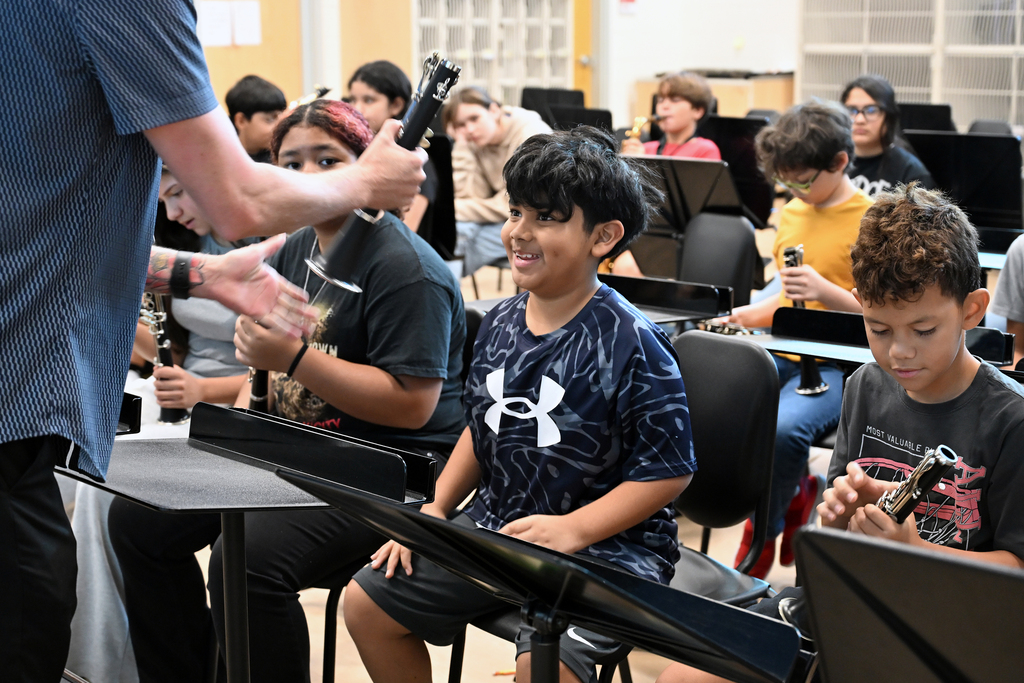 An adult male shows a clarinet to a boy in a classroom. Other children sit in chairs, playing flutes.