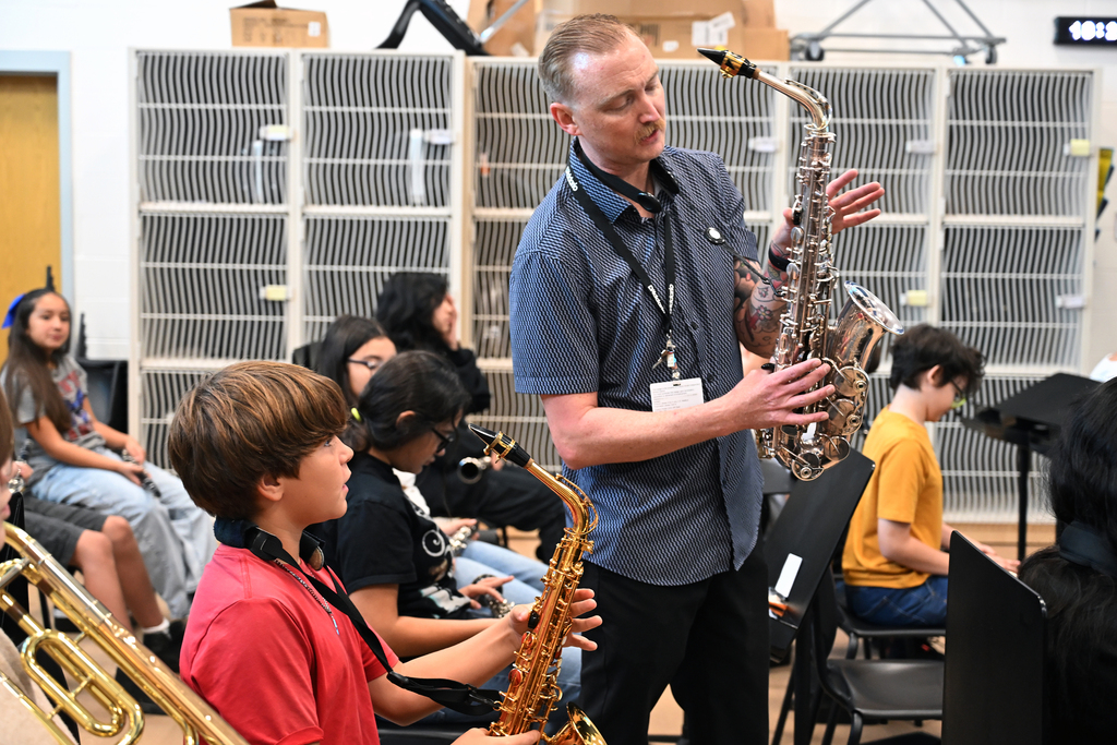 A man demonstrates a saxophone to a group of children seated in a room. Some students play saxophones.