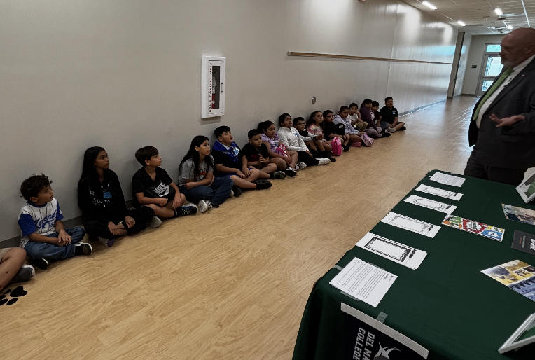 A row of children sits on a floor. A man stands near a table with papers and a frame on the wall.