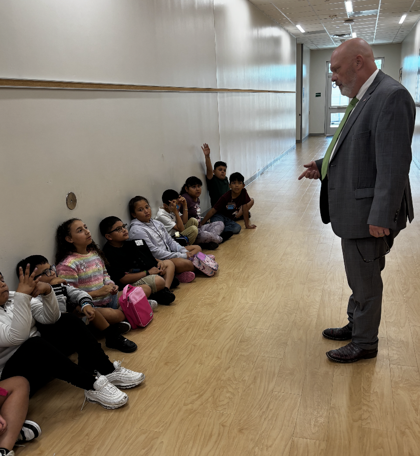 A man in a suit and tie speaks to children sitting in a line against a wall.