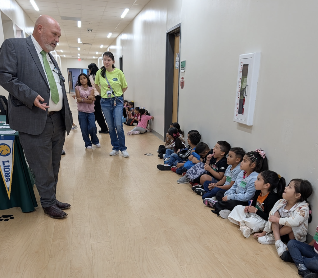 A man in a suit stands before a seated line of children in a school corridor. Other adults are in the background.