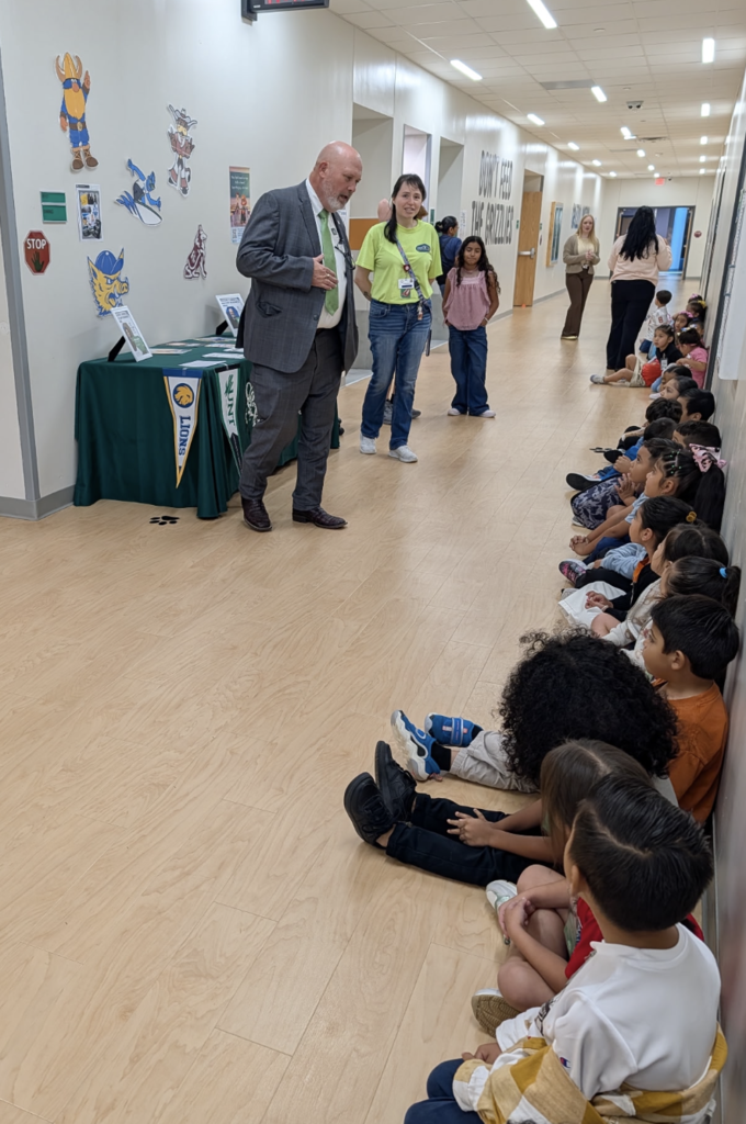 A hallway with wooden floors; a man in a suit speaks to children seated against the wall.