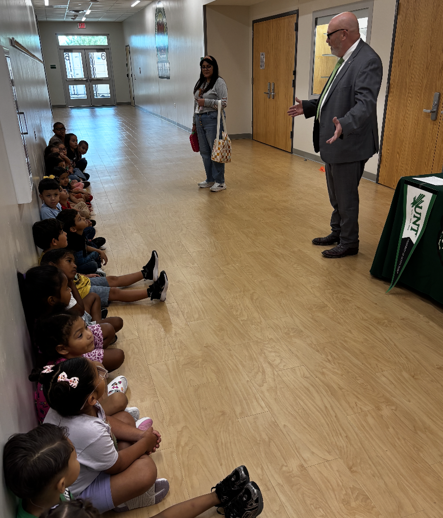 A group of children sits in a hallway, listening to a man in a suit standing. A woman stands nearby.