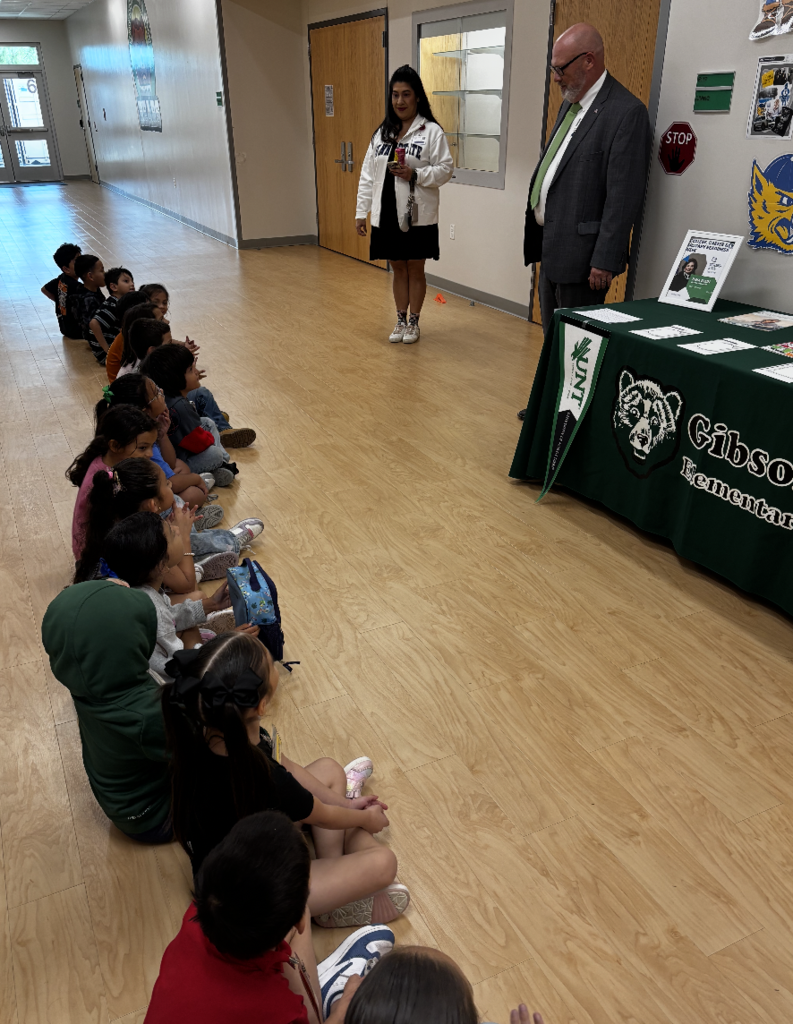 Adults and children sit in a row. A woman stands before them. A table displays a green banner.