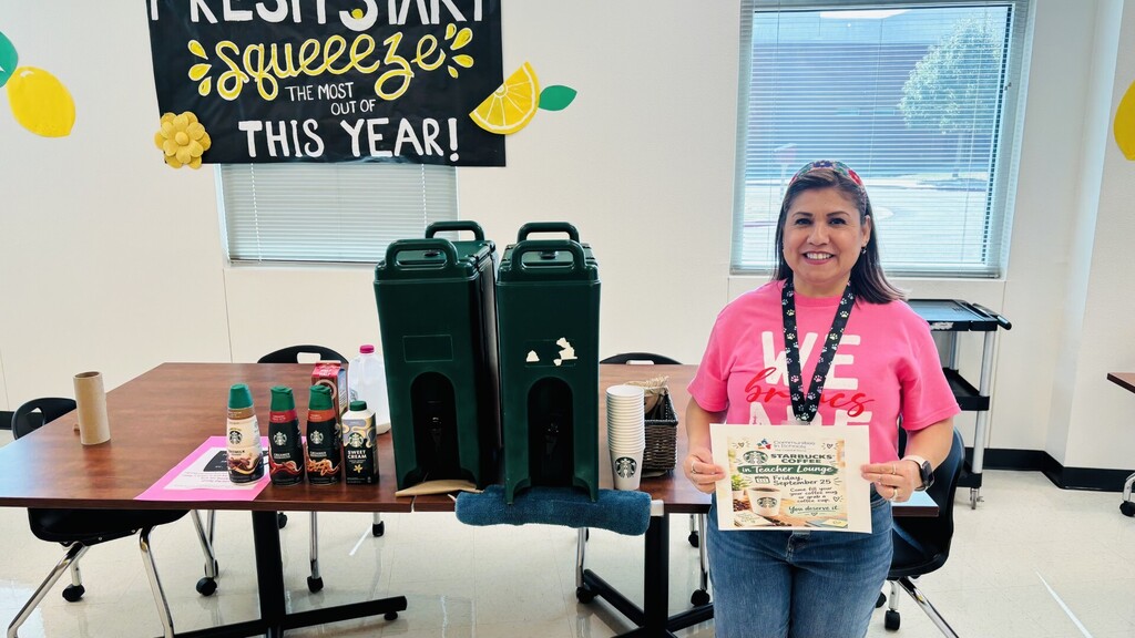 A woman in a pink shirt holds a certificate in a room with a table, chairs, and two green dispensers.