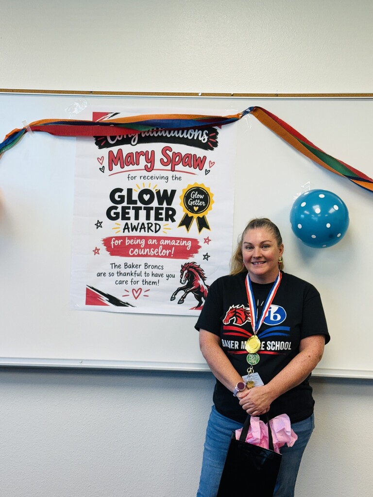 A woman wearing a black T-shirt, jeans, and a necklace stands next to a whiteboard. Behind her is a colorful banner and a poster for a Glow Getter Award.