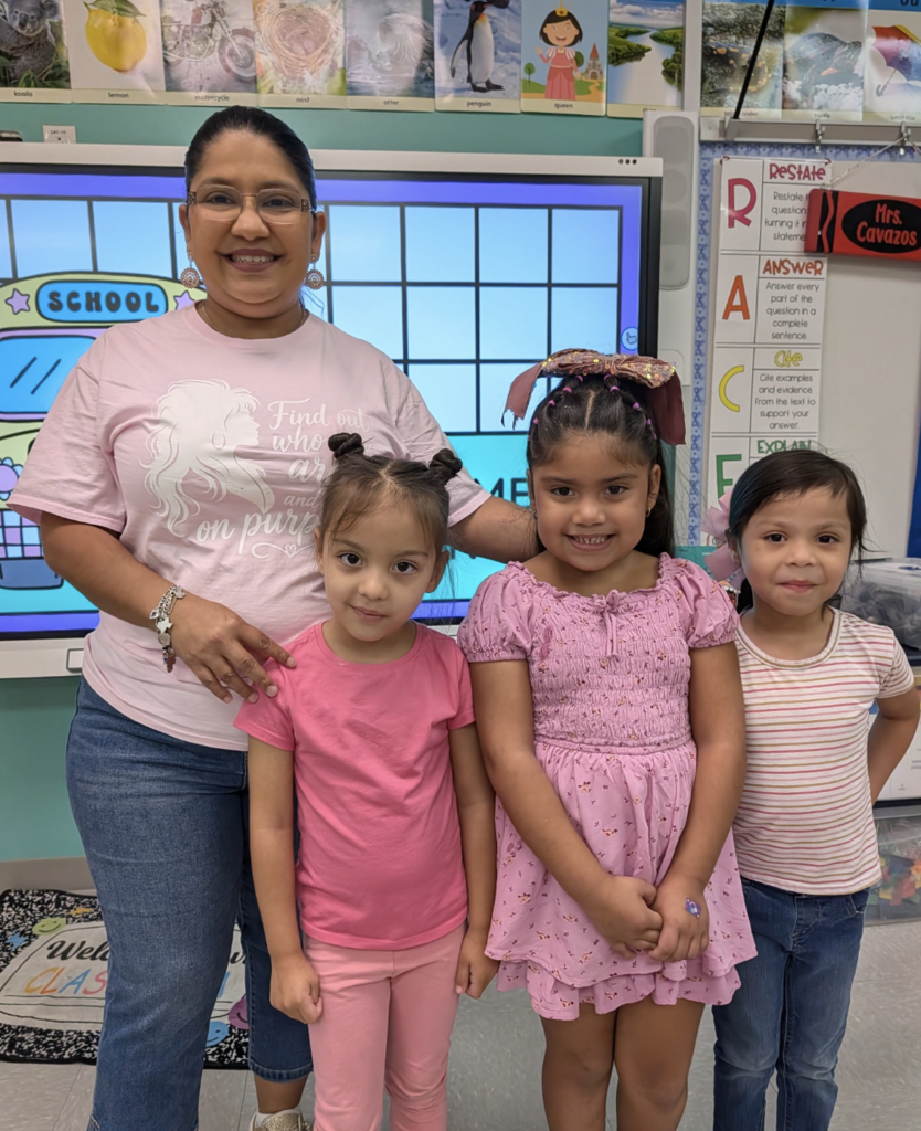 Woman with three girls in front of a classroom screen. The woman wears a pink shirt with writing.