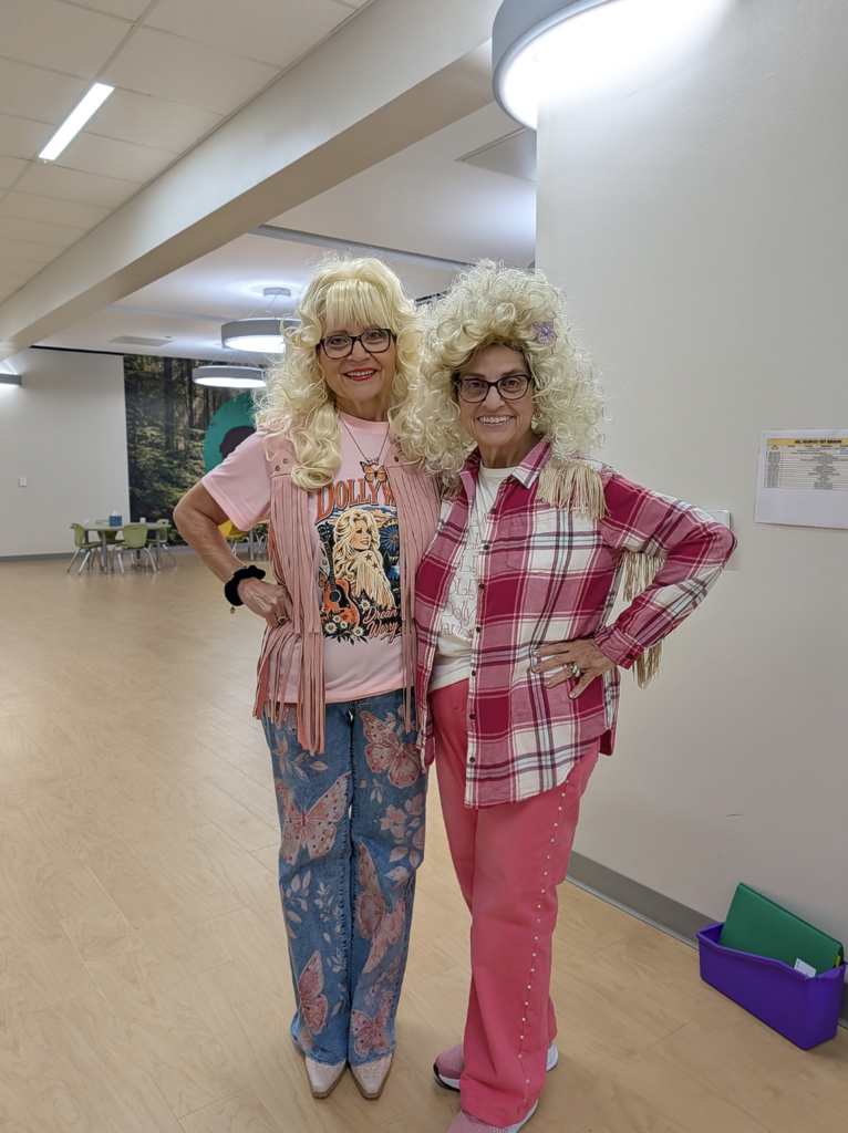 Two women with blonde wigs and glasses, wearing pink pants, standing together in a hallway.