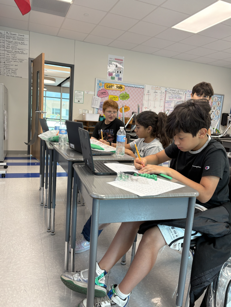 Children seated at desks in a classroom, one child with a pen, another with a laptop, all wearing casual clothes.