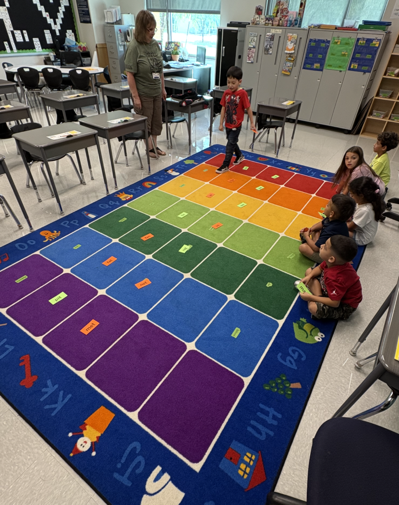 A classroom with a colorful carpet floor with squares, children sitting, and a teacher standing nearby.