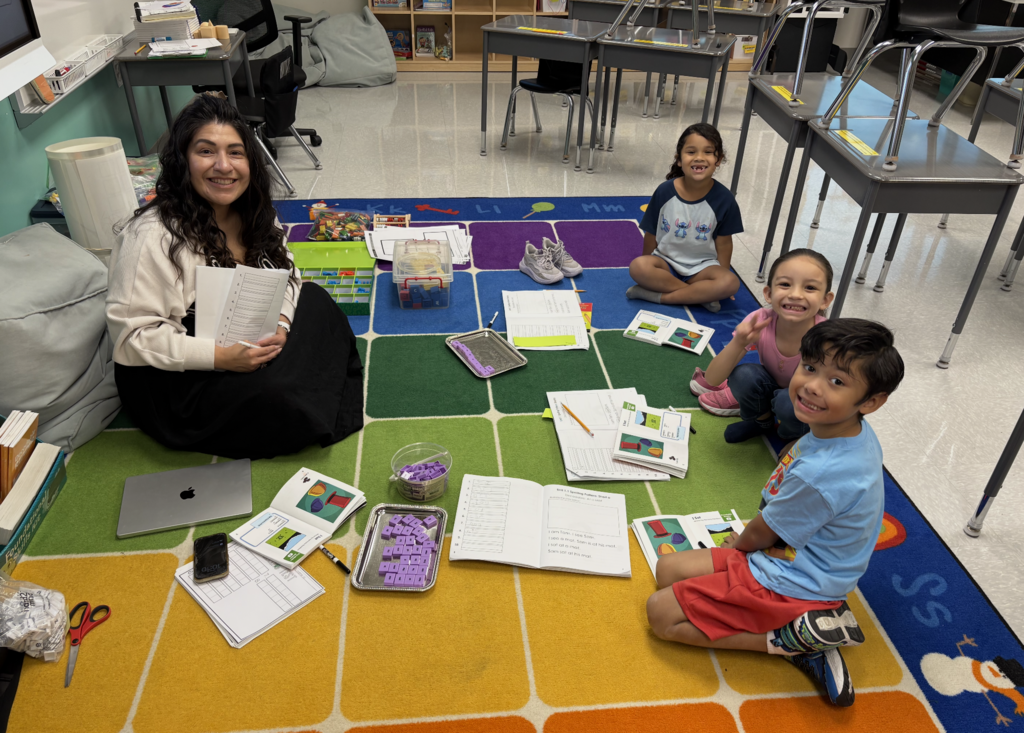 A woman and three children sit on a multicolored rug in a classroom. Papers, scissors, and books are scattered around.
