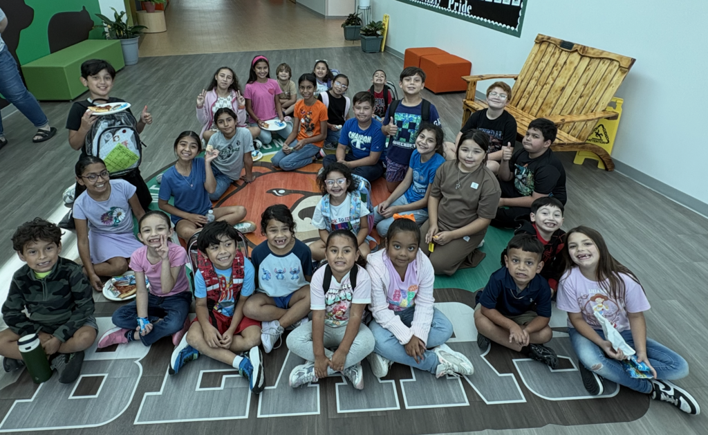 A group of children in a classroom. Some sit on a basketball court rug; some are standing.