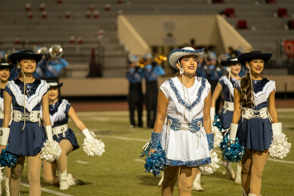 Cheerleaders in blue uniforms and cowboy hats perform on a football field, with a band in the background.