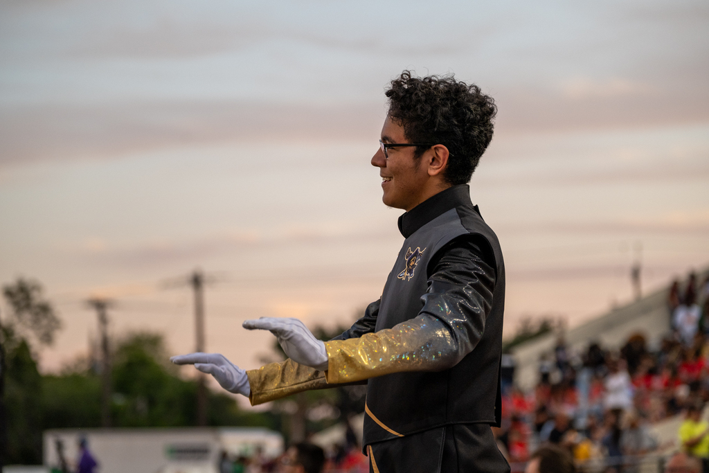 A person conducts with one hand while wearing a black and gold jacket. An audience is seated in the background.