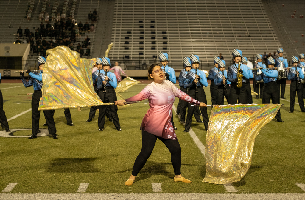 Marching band on a field with a woman holding gold flags, others behind her in blue uniforms, stadium in background.