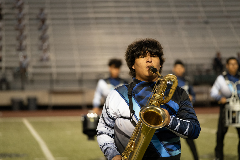 A saxophone player stands on a football field, surrounded by other musicians playing drums in a stadium.
