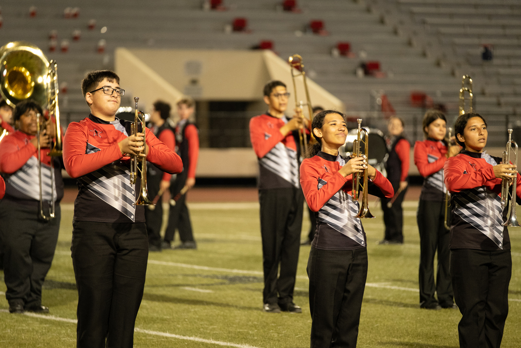 A group of students in matching uniforms play musical instruments on a field, with bleachers in the background.
