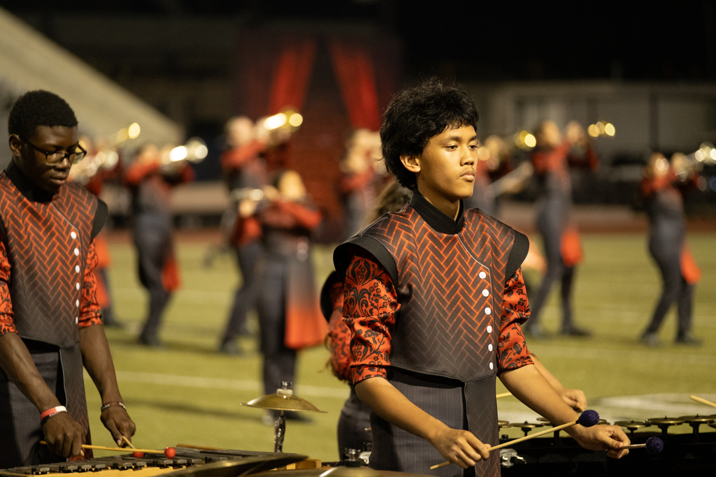 Musicians play xylophones on a football field with a marching band in red and black uniforms.