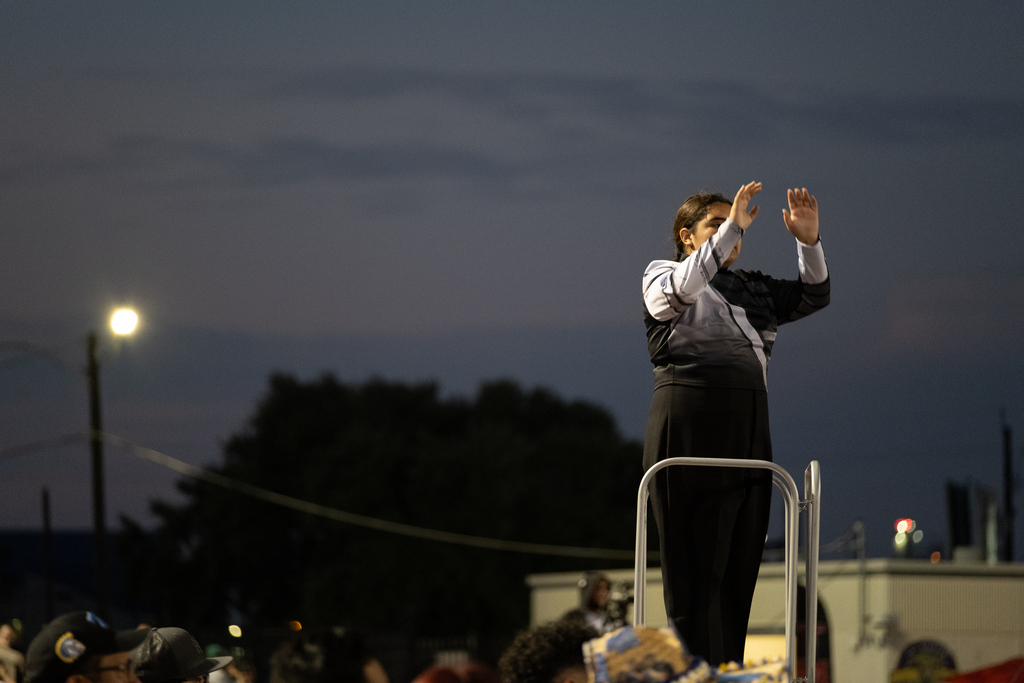 Person standing on elevated platform with hands raised, wearing dark clothing, performing in front of audience at night.
