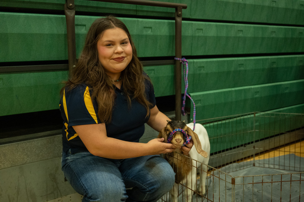 Woman kneeling holding a goat in a cage, in a room with green bleachers.