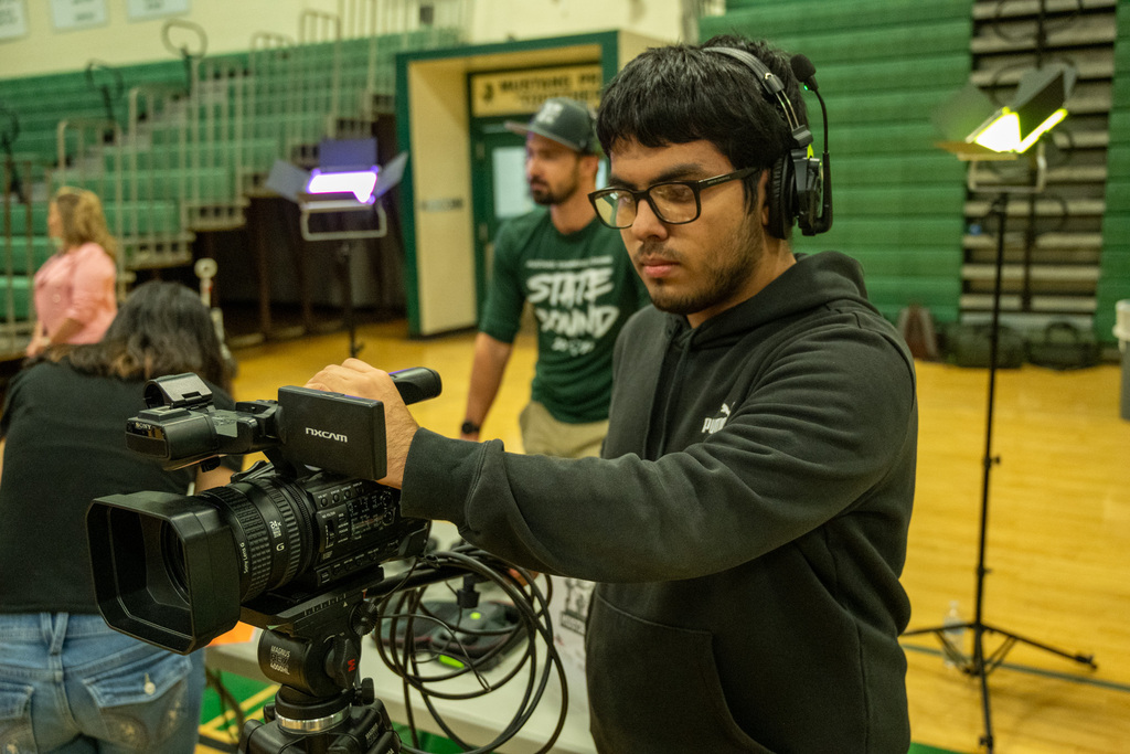 A man in a black hoodie operates a video camera in a gym with a green wall, while others look on.
