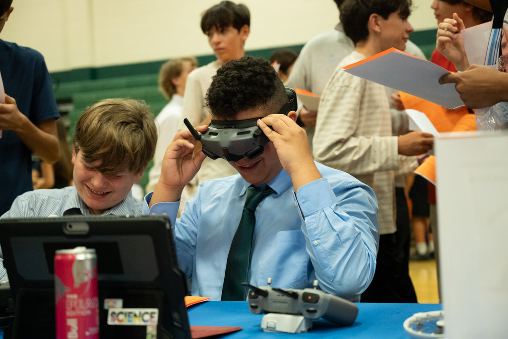 Two boys sit at a table wearing VR goggles, one holding a remote. Behind them, people stand or sit, some holding papers.