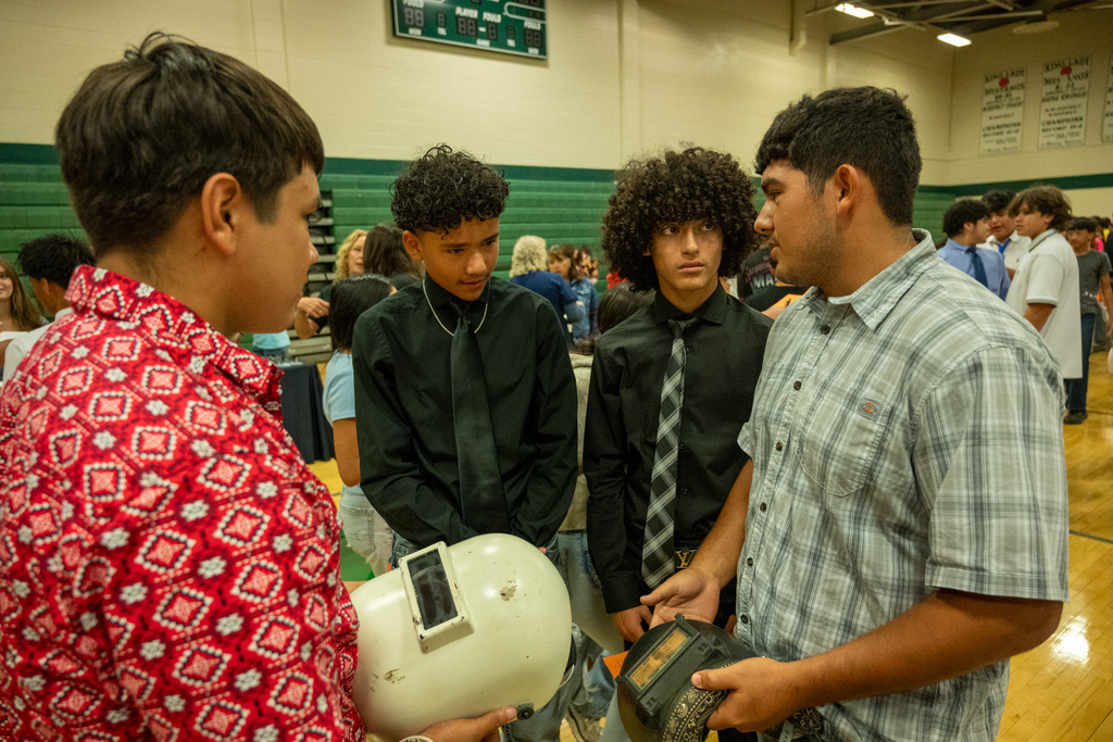 Four boys are standing in a gym. One holds a welding helmet, while others look at items in their hands.