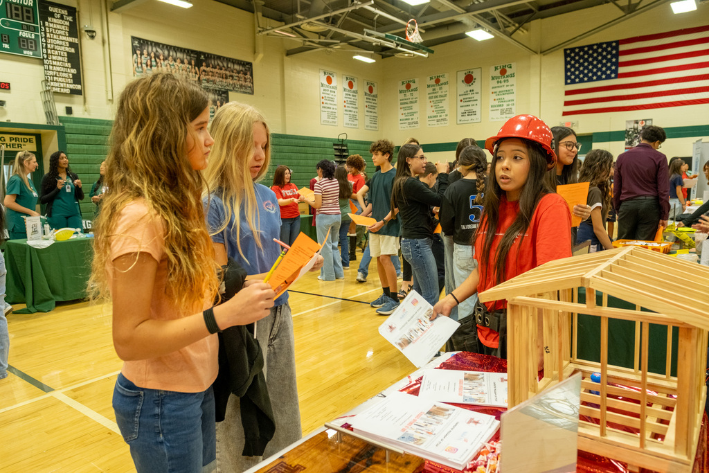 Four students examine a birdhouse model at a school event, with a basketball court, green tables, and a flag in the background.