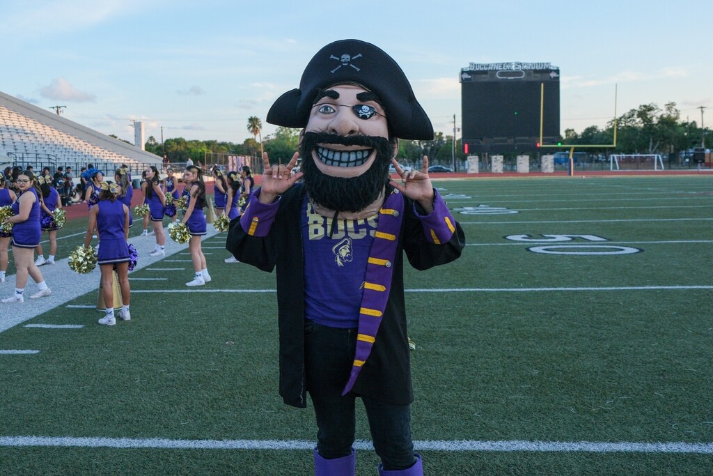 Mascot with pirate hat, purple and black outfit, and beard on a football field. Cheerleaders in purple and white uniforms in background.