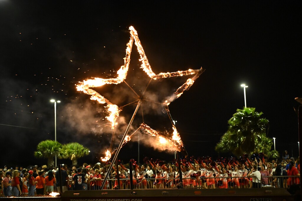 A burning star shape in a night sky. Trees and street lights surround the crowd watching.