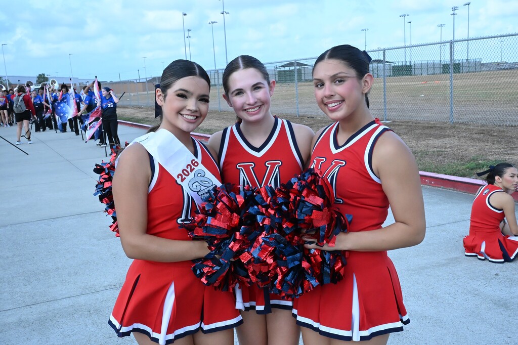 Three cheerleaders in red uniforms with white trims stand together, one holding pom-poms. Behind them, a crowd gathers, and a metal fence surrounds the area.