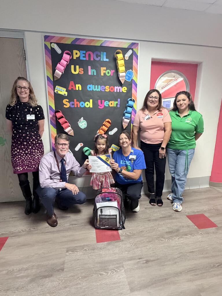 Six adults and a child pose behind a chalkboard with "Pencil Us In For An Awesome School Year" and a pink door.