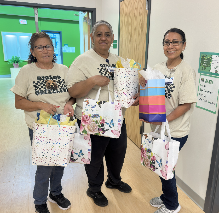 Three women stand together holding gift bags. They wear matching white T-shirts with text and designs.
