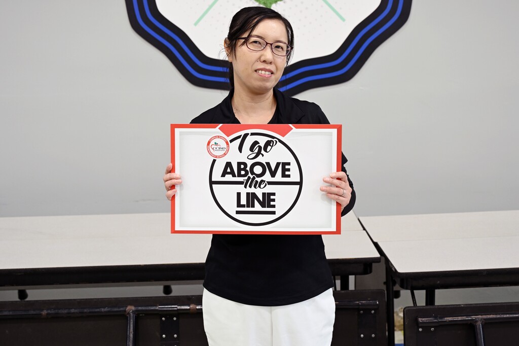 Woman in glasses holds a sign that reads 'I Go Above the Line' in front of tables.
