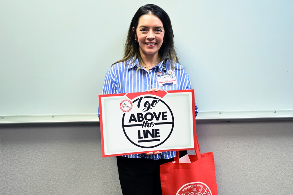 A woman holds a sign saying "To Above the Line" in front of a whiteboard.