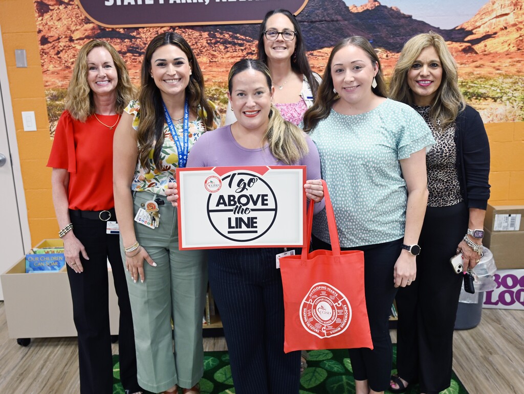 Five women pose with a sign in a room with desert mural. Woman in center holds a red tote.