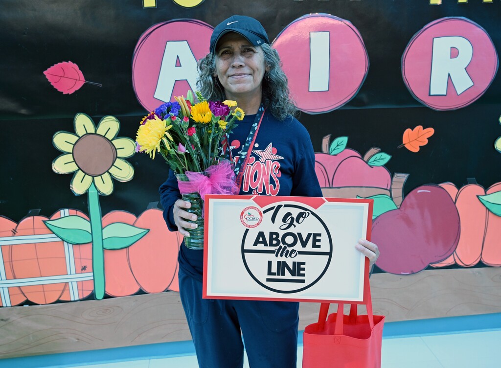 Woman holds a sign with text, flowers, and a red bag. The background includes painted pumpkins, flowers, and a banner.
