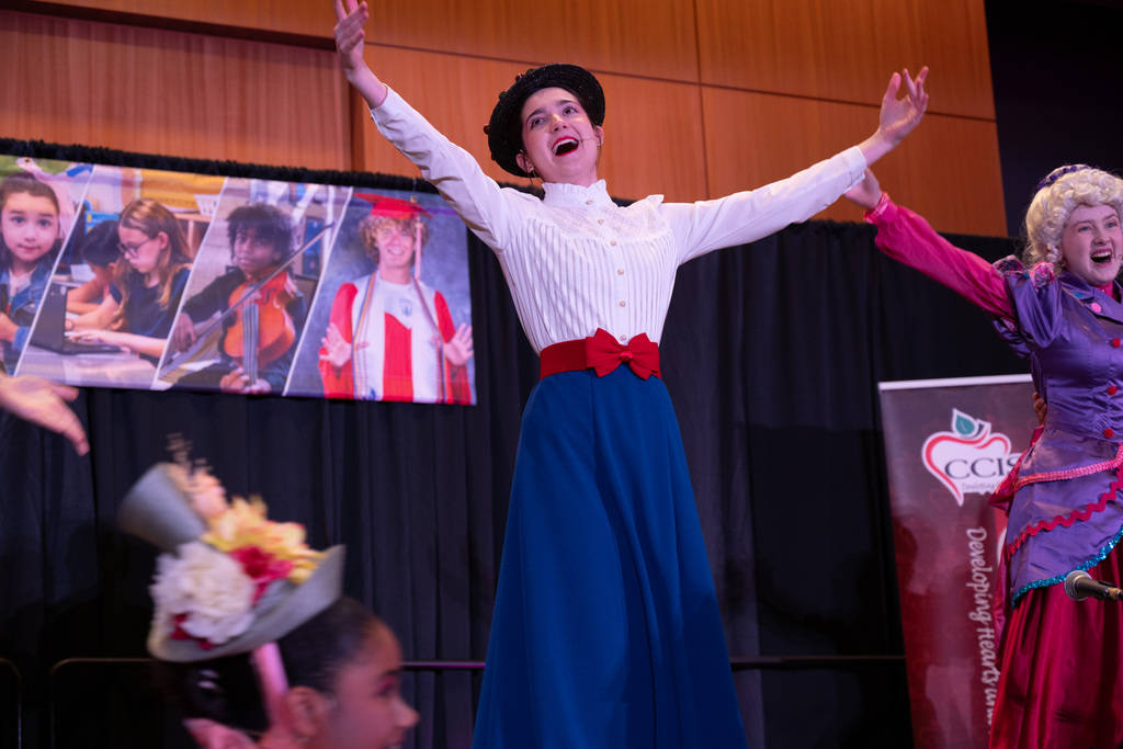 Performers on stage, one in a white blouse and blue skirt, another in purple and pink. Background with photos.