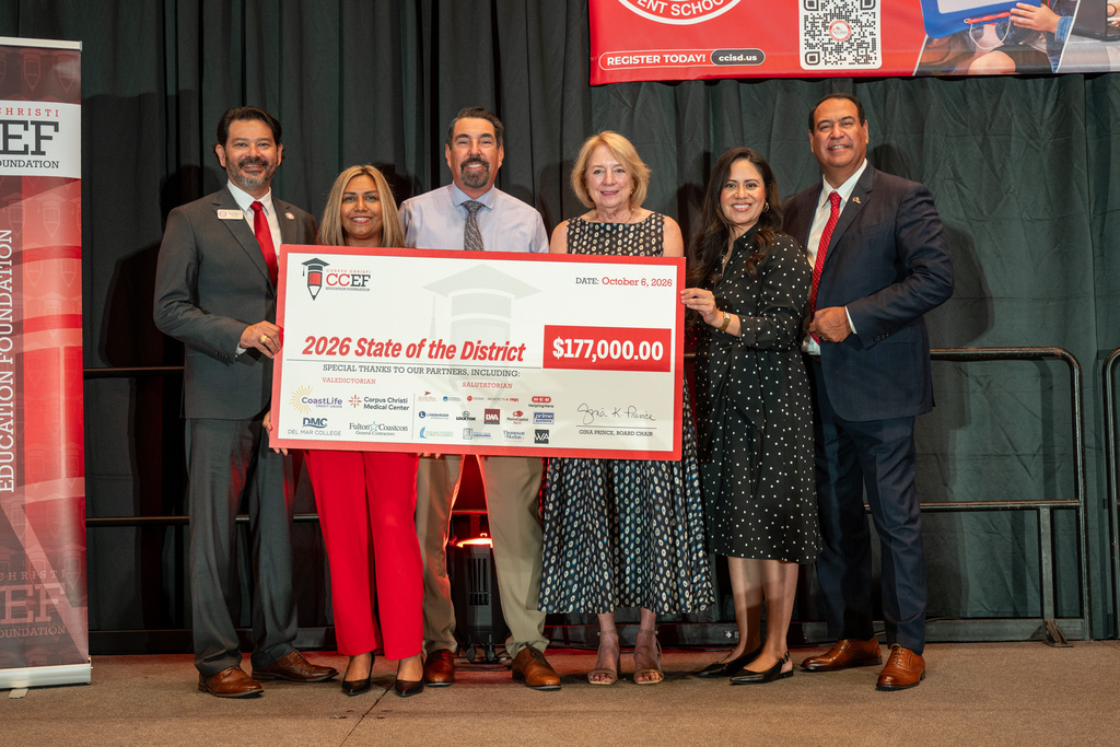 Six adults stand on stage holding a large check. Two are in red suits, one in red pants, and three in black dresses.