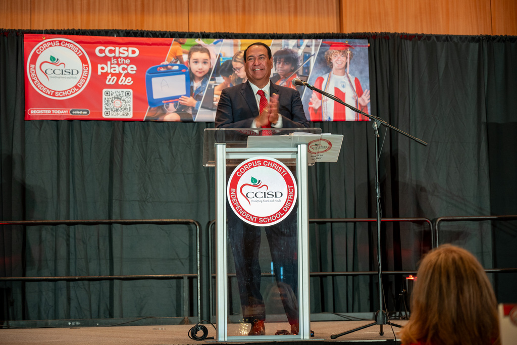 A man speaks at a podium with a microphone in front of a banner and audience.