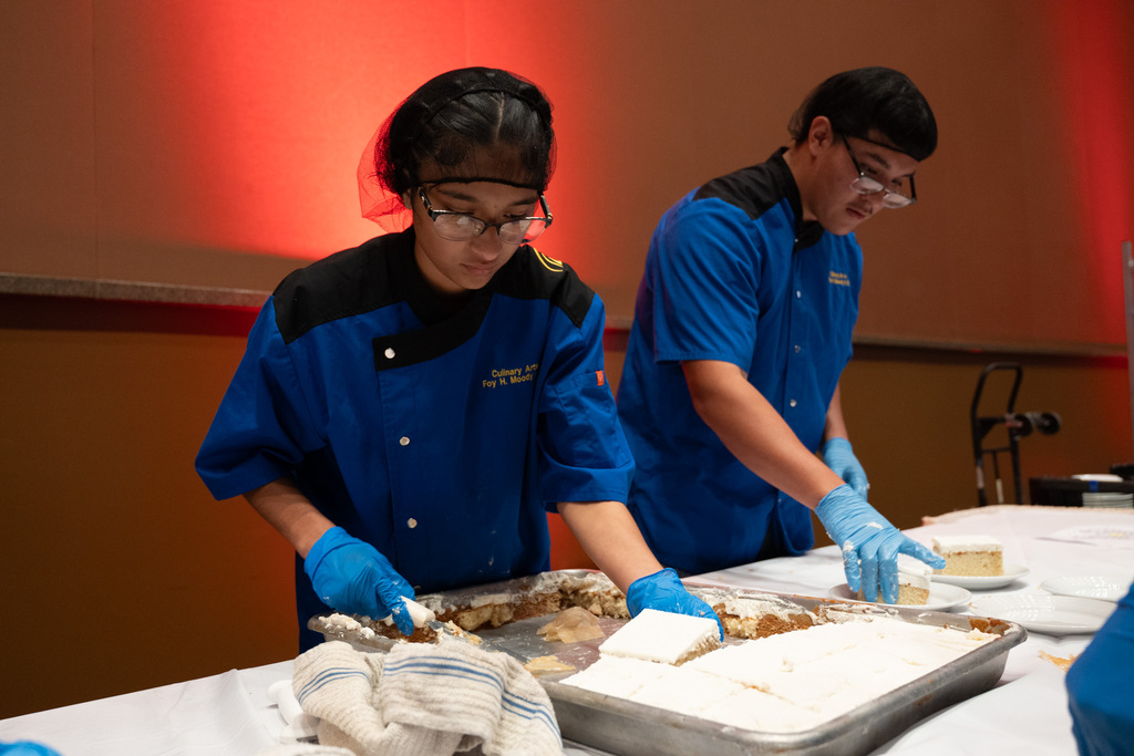 Two individuals in blue uniforms and gloves are preparing food on a table with a large tray.
