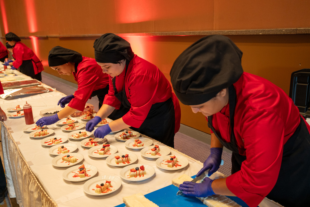 Four chefs in red uniforms prepare food on a long table with multiple plates and a blue mat.