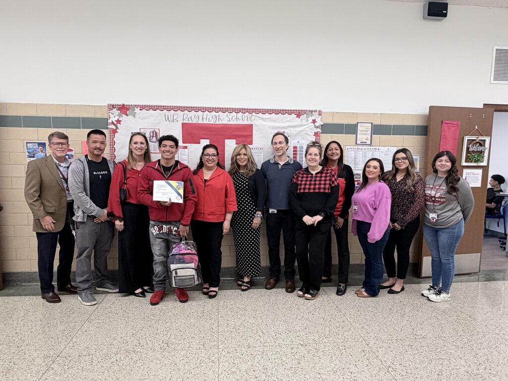 Eleven people stand in a line for a group photo in a room with white walls and a bulletin board.