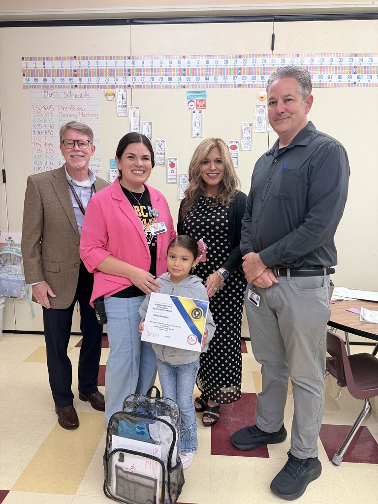 Four adults and a child stand in a classroom with a girl holding a certificate. Behind them, papers are pinned to a bulletin board.