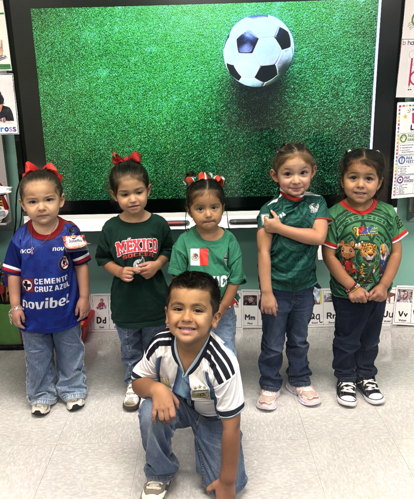 Group of children posing for a picture in front of a large screen with a soccer ball.